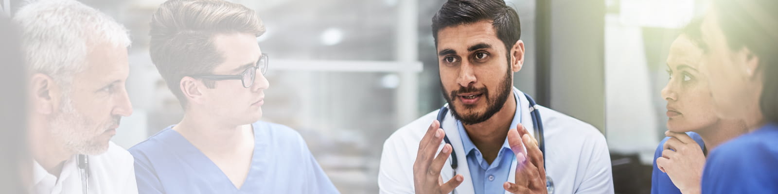 Physician in white coat talking with four listening clinicians in blue scrubs.