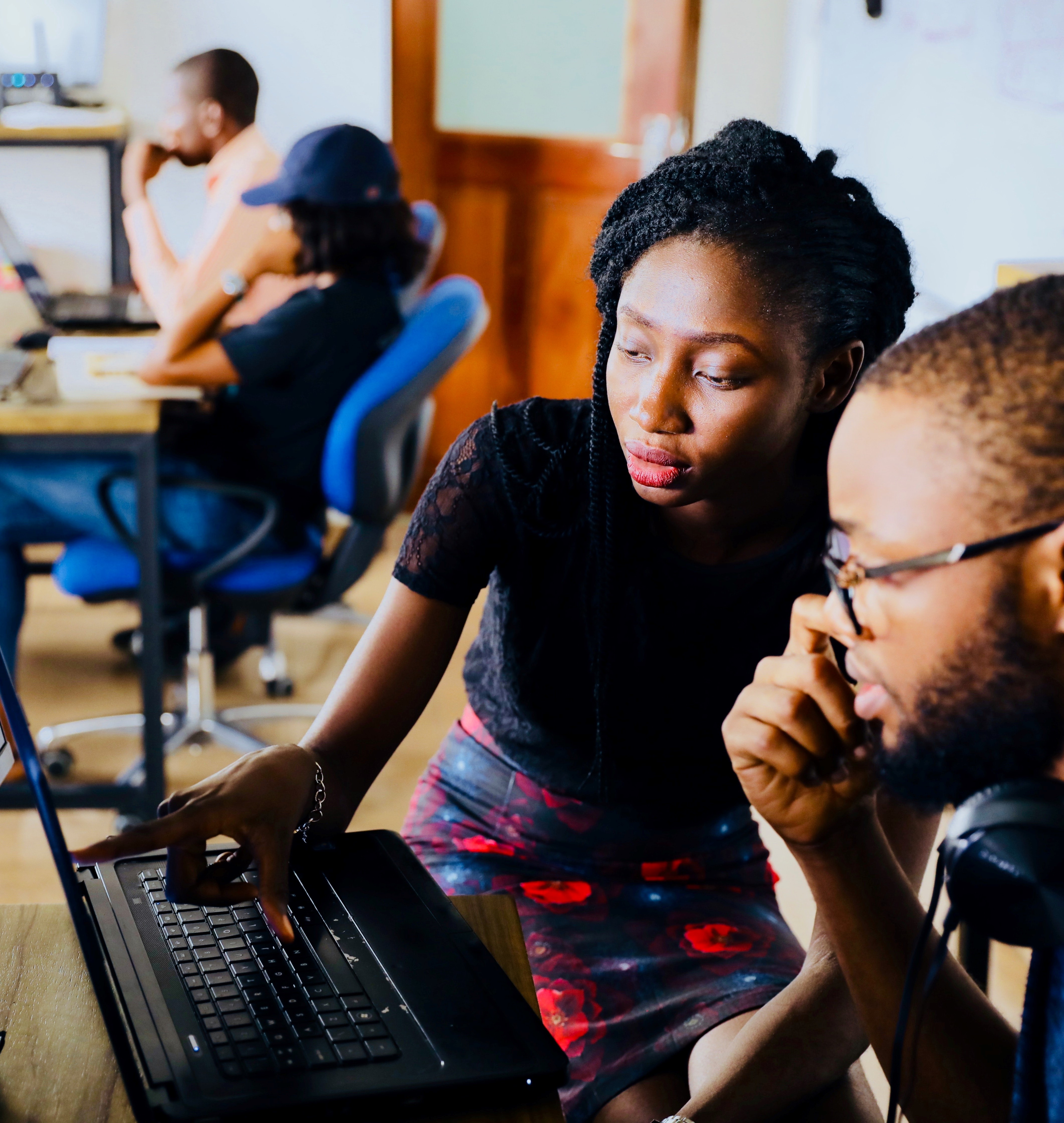 Woman guiding a man by pointing at a laptop screen in a classroom setting.